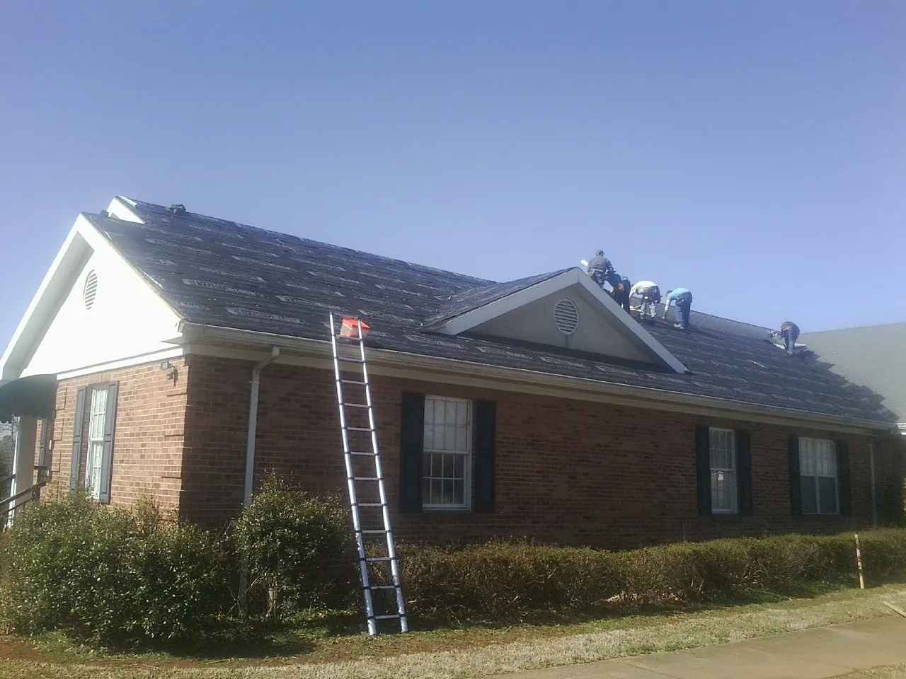 Crew tearing shingles off a brick ranch roof with a ladder set — Metal Roof Repair work by our crew in Sutherland