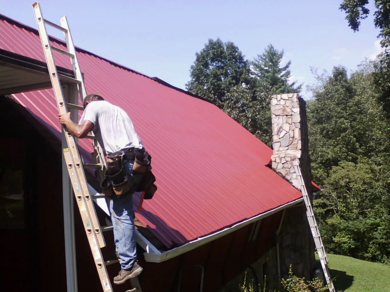 Roofer on a ladder beside a red metal roof and stone chimney — Soffit & Fascia Repair by Roofing Brothers in Sutherland